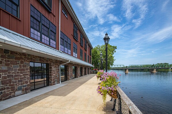 A building with a stone facade and large windows, a concrete walkway, and a river with a bridge in the background. - Ghost Light Inn (New Hope)
