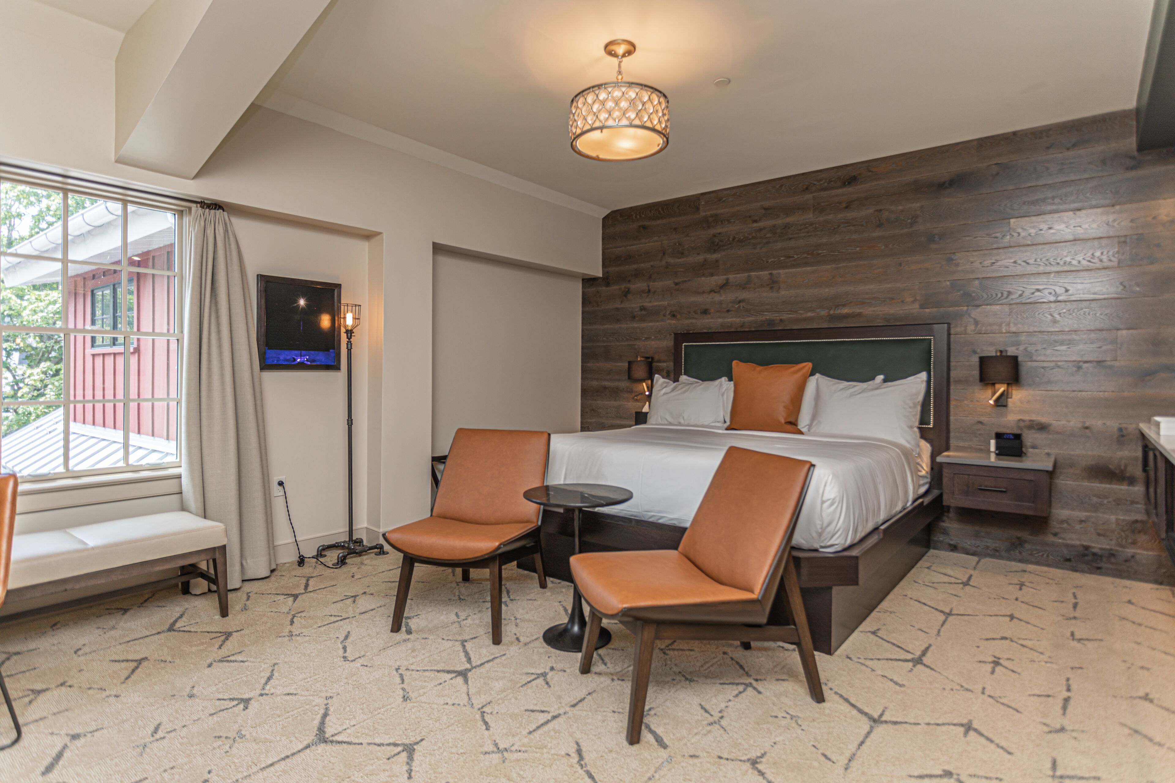 A hotel room with a large bed, two chairs, a small table, a TV, and a window with curtains.
