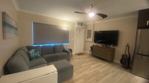 A living room with a grey sofa, a flat-screen TV, a ceiling fan, and a refrigerator. - Sudio Apt Right on the Beach! (Lauderdale by the Sea)