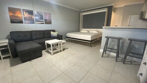 Un espace de vie compact comprenant un canapé, une petite table blanche, un lit et un coin cuisine.