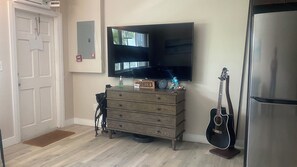Una habitación con una televisión de pantalla plana montada en la pared, un aparador de madera con cajones, una guitarra y un refrigerador.