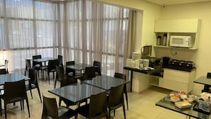 Zona pranzo con tavoli e sedie, cucina con elettrodomestici e vista attraverso tende leggere.