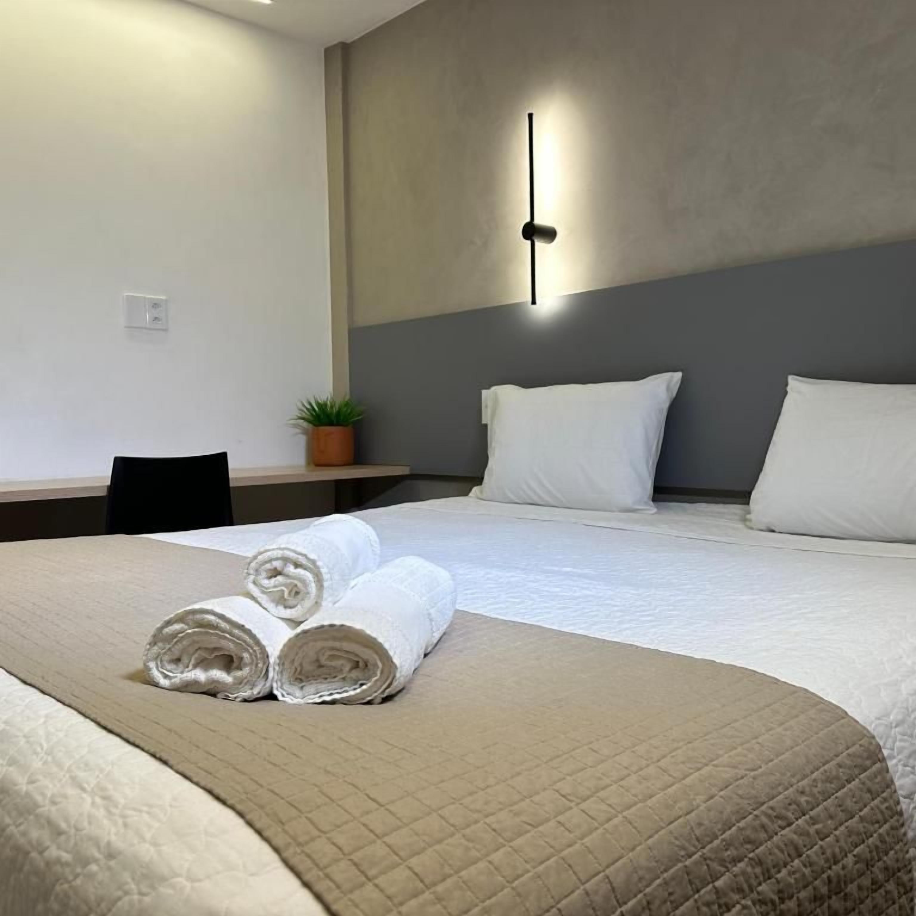 A neatly made bed with rolled towels and a beige quilted blanket, a small potted plant on a nightstand, and a wall-mounted light fixture.