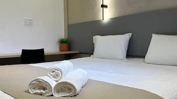 A neatly made bed with rolled towels and a beige quilted blanket, a small potted plant on a nightstand, and a wall-mounted light fixture.