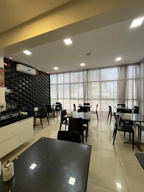Un comedor moderno con mesas y sillas, una encimera negra con electrodomésticos de cocina y un amplio ventanal con cortinas.