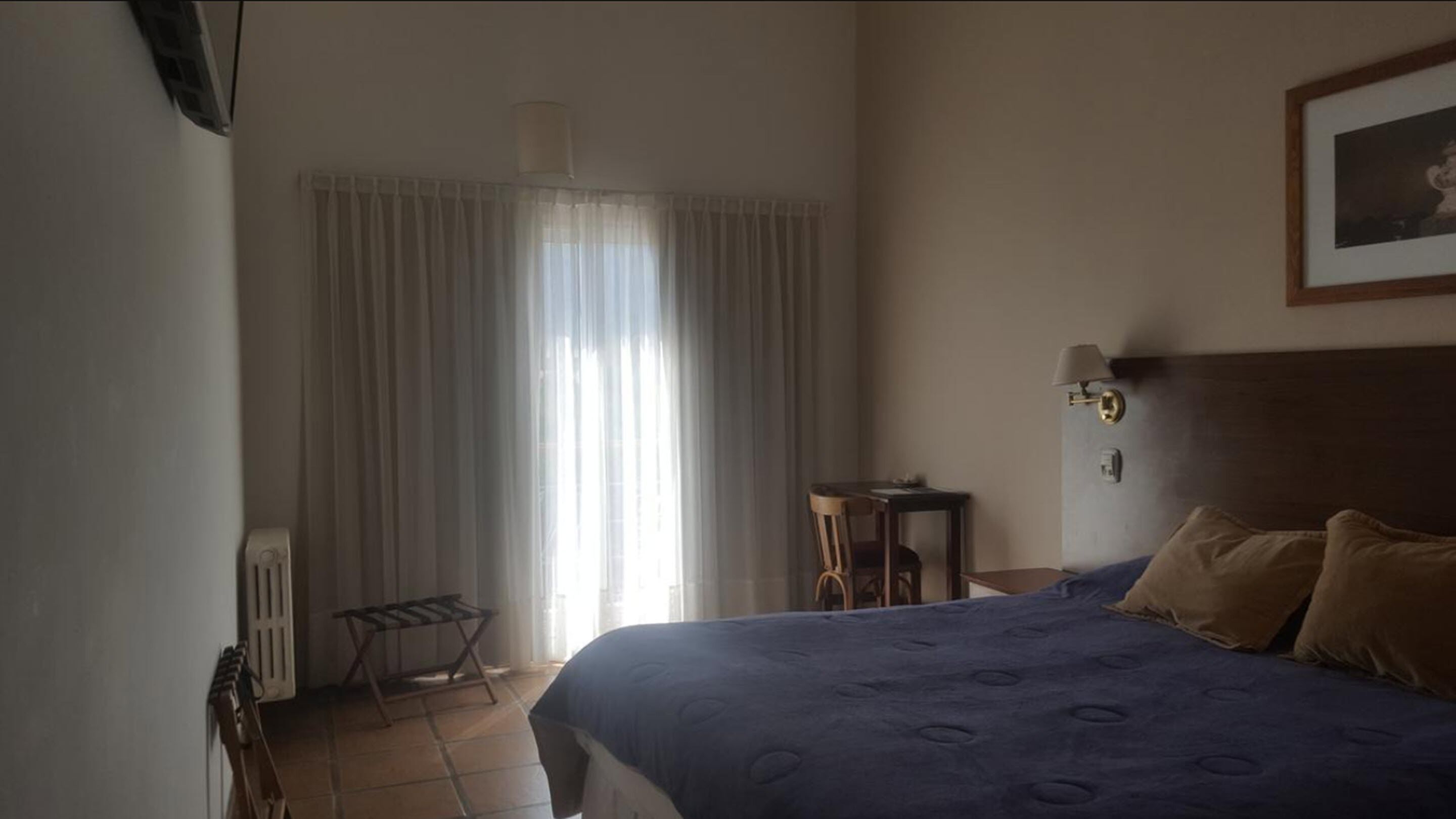 Een hotelkamer met een bed, een klein tafeltje, een stoel, een radiator en een raam met gordijnen.