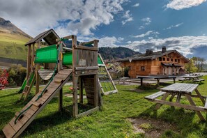 A wooden play structure with a slide and climbing area, picnic tables, and a chalet-style building in the background.