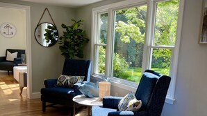 A cozy living room with two dark blue armchairs, a round coffee table, and a large window offering a view of a lush garden.