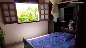 A bedroom with a bed, a window with curtains, a bedside table with a lamp, and a shelf with decorative items. - TITIRI apartment with terrace and garden (Sainte-Anne)