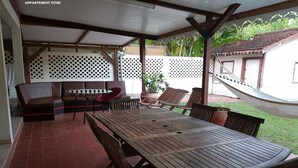 A covered patio area with a wooden table and chairs, a sofa, and a hammock. - TITIRI apartment with terrace and garden (Sainte-Anne)