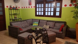 A living room with a sectional sofa, a glass coffee table, and a black metal chair. There is a window with wooden frames and a wall-mounted fan. - TITIRI apartment with terrace and garden (Sainte-Anne)
