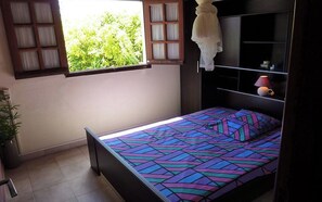 Une chambre à coucher avec un lit, une fenêtre avec des rideaux, une table de chevet avec une lampe et une étagère avec des objets décoratifs.