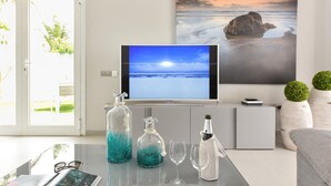 Un saló modern amb una televisió que mostra una escena de platja, una taula de vidre amb objectes decoratius i un quadre de platja a la paret.