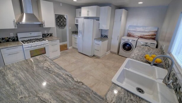Una cocina moderna con armarios blancos, una campana extractora de acero inoxidable, un horno empotrado, un refrigerador, una lavadora y un lavamanos con encimera de mármol.