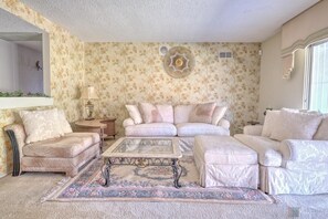 Sala de estar com papel de parede floral, um sofá bege, mesa de centro de vidro e tapete estampado.