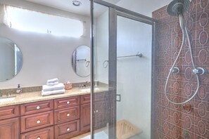 A bathroom with a glass-enclosed shower, wooden vanity with drawers, a round mirror, and a tiled backsplash.