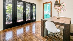 A room with a wooden floor, a dining table with chairs, a vase with flowers, and a view through a glass door to an outdoor area with a chair and greenery. - Oak Haven (Altadena)