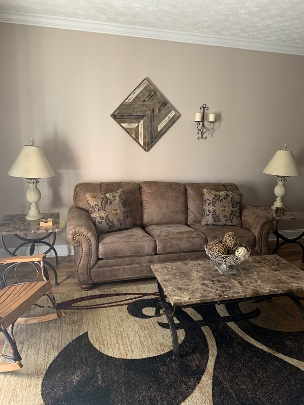 Een woonkamer met een bruine bank, twee bijzettafels met lampen, een salontafel met decoratieve objecten, een kamerkleed met een patroon en een geometrisch kunstwerk aan de muur.