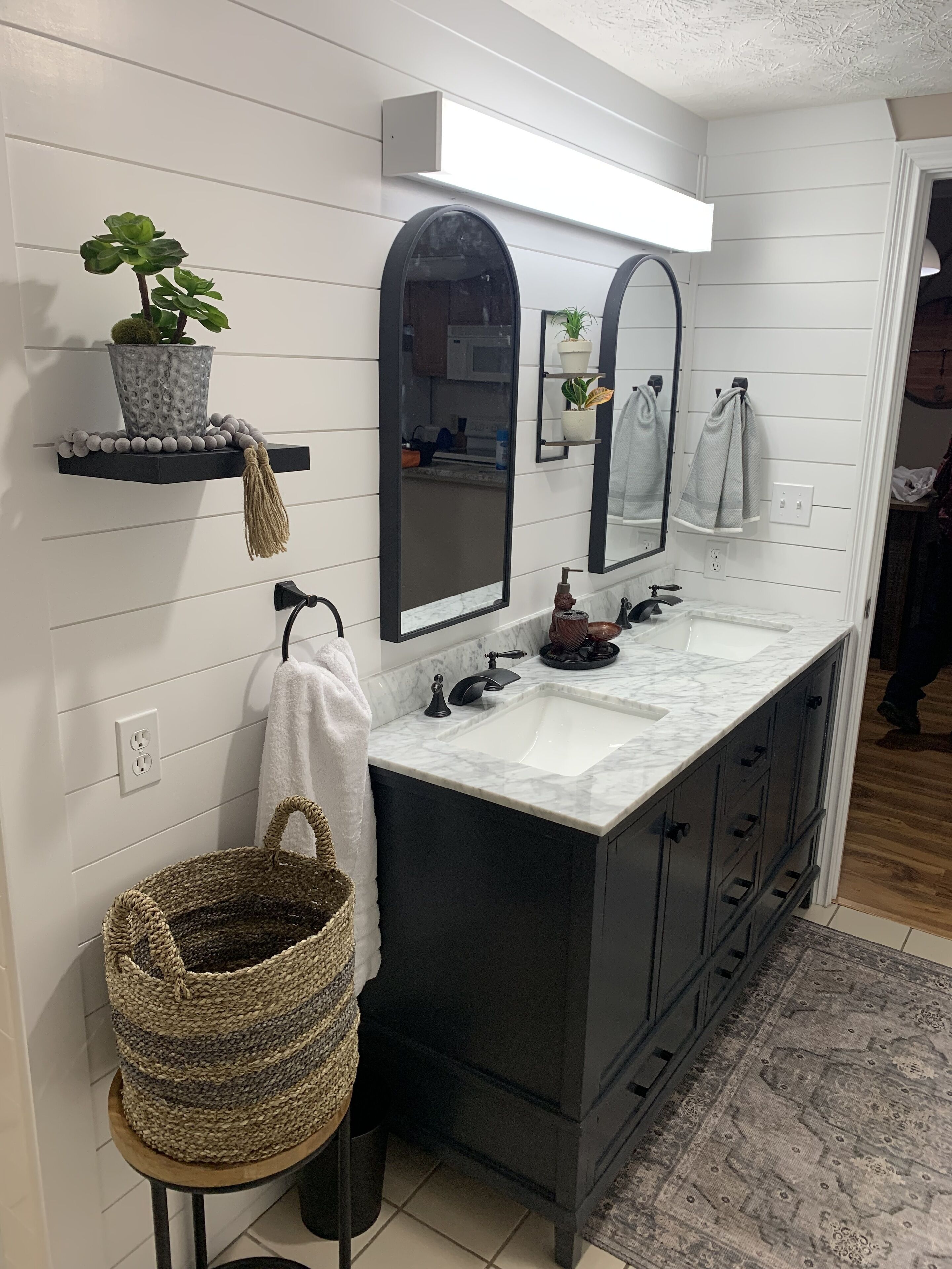Un baño moderno con pared de azulejos tipo subway blancos, un mueble de tocador negro con lavamanos blanco, un espejo grande y una planta en una repisa flotante.