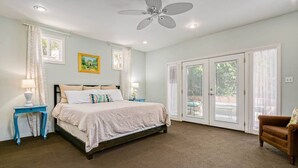 A bedroom with a bed, a nightstand, a chair, a ceiling fan, and a large window with a view of the outdoors.