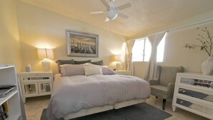 A bedroom with a bed, bedside tables, a bookshelf, a chair, a ceiling fan, and a window with curtains.