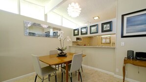 A dining area with a wooden table and four chairs, a decorative mirror, framed pictures on the wall, and a small kitchen area with a countertop and sink.