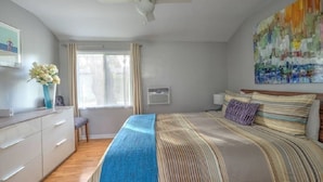 A bedroom with a bed, a ceiling fan, a window with curtains, a bedside table with a vase of flowers, and a colorful abstract painting on the wall. - The Willows (Pasadena)