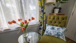 A room with a glass table holding a vase of tulips, a tufted green armchair with floral cushions, and a window with sheer curtains. - The Sycamore (Pasadena)