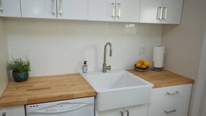 A kitchen with white cabinets, a wooden countertop, a built-in dishwasher, a sink, and a potted plant. - The Sycamore (Pasadena)