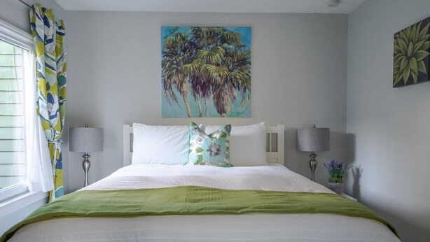 A neatly made bed with white linens and a green throw blanket, flanked by two bedside lamps and a nightstand with a lamp. A tropical-themed painting adorns the wall above the bed. - The Sycamore (Pasadena)