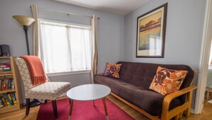 A living room with a sofa, a chair, a small table, a bookshelf, and a window with curtains. - The Deodora (Pasadena)