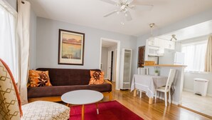 A living room with a brown sofa, a white table, and a red rug. There is a kitchen area with white cabinets and a dining table. - The Deodora (Pasadena)