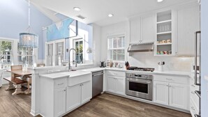 A modern kitchen with white cabinetry, a central island, stainless steel appliances, and a dining area with a wooden table and chairs. - Luxury Watersound Getaway with Golf Cart (Watersound)