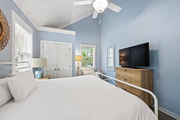 A bedroom with a bed, a TV, a ceiling fan, and a window with blinds.