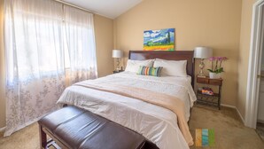 A bedroom with a bed, a bench, a nightstand, a lamp, and a painting on the wall. - The Madison (Pasadena)