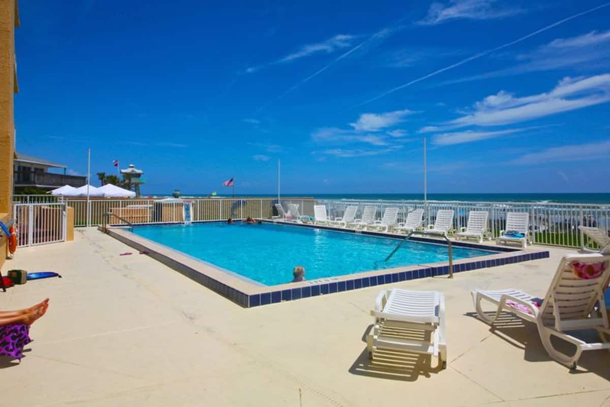 A rectangular pool with a tiled edge, surrounded by a white fence and white lounge chairs.