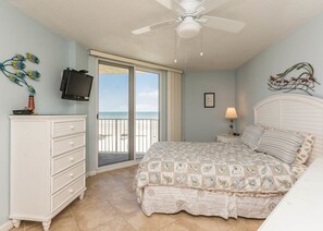 Une chambre à coucher comprenant un lit, une commode, un téléviseur, une lampe et une vue sur la plage, accessible par une porte coulissante.