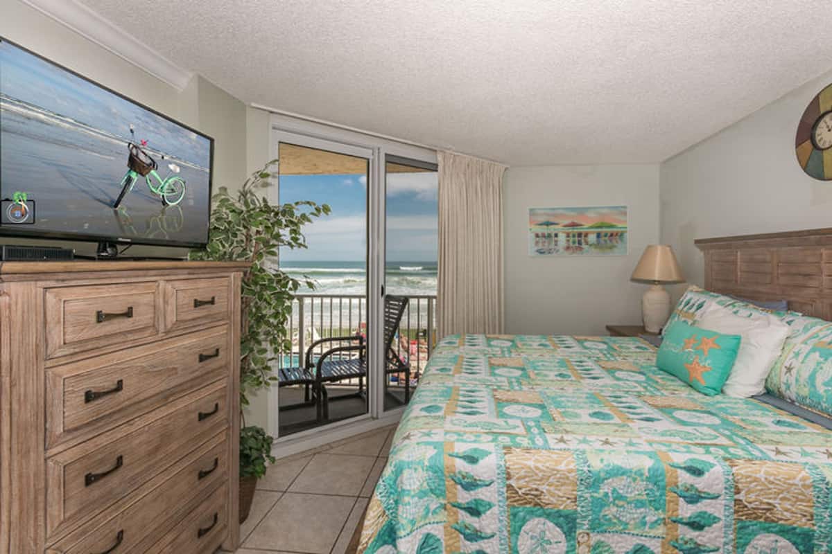 A bedroom with a bed, dresser, TV, balcony with a view of the ocean, and a painting on the wall.