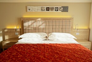 A neatly made bed with a plaid headboard, white pillows, and a red patterned bedspread. There are two bedside tables with lamps and a wall-mounted shelf above the bed. - Hotel Alpina (Pinzolo)