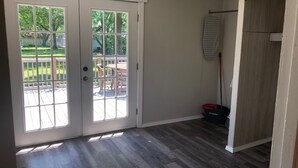 Una habitación con piso de madera, una puerta de vidrio corredera que da a un patio exterior con mesa y sillas, y una escoba apoyada en la pared.
