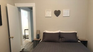 Un dormitorio con cama, dos mesitas de noche, una mesita auxiliar, una puerta y un pasillo que conduce a otra habitación.