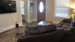 Una sala de estar con un sofá de cuero, un televisor de pantalla plana montado en la pared y una mesa de comedor de madera.