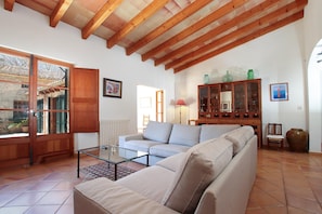 Un salon avec un canapé, une table basse en verre, des poutres en bois au plafond, un meuble et une vue sur l’extérieur, visible à travers une porte.