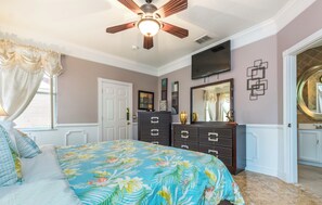 Een slaapkamer met een bed, een dressoir, een plafondventilator, een televisie en een badkamers deur.