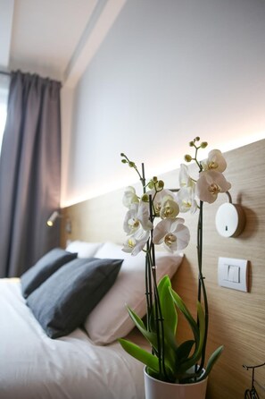 A hotel room with a bed, white pillows, a bedside table with a white vase containing white orchids, and a wall-mounted light switch. - Bed and Bike Verona (Verona)