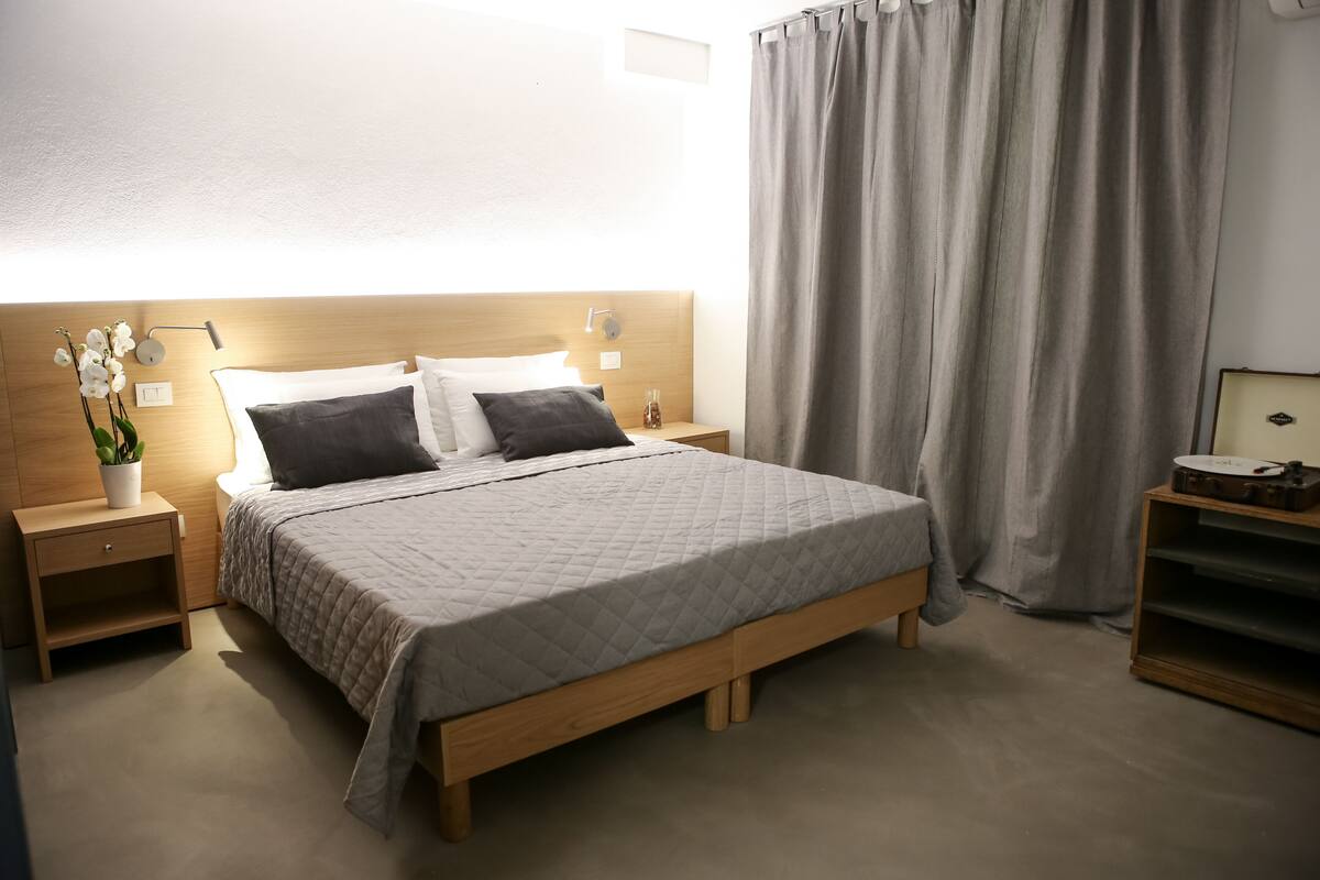 Un lit bien fait, avec une couverture matelassée, une tête de lit en bois, des tables de chevet avec des lampes, une plante en pot et un rideau.