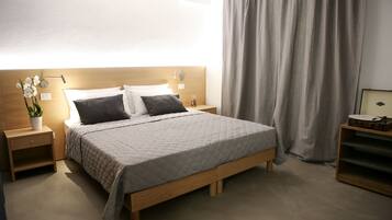 A neatly made bed with a quilted cover, wooden headboard, bedside tables with lamps, a potted plant, and a curtain.