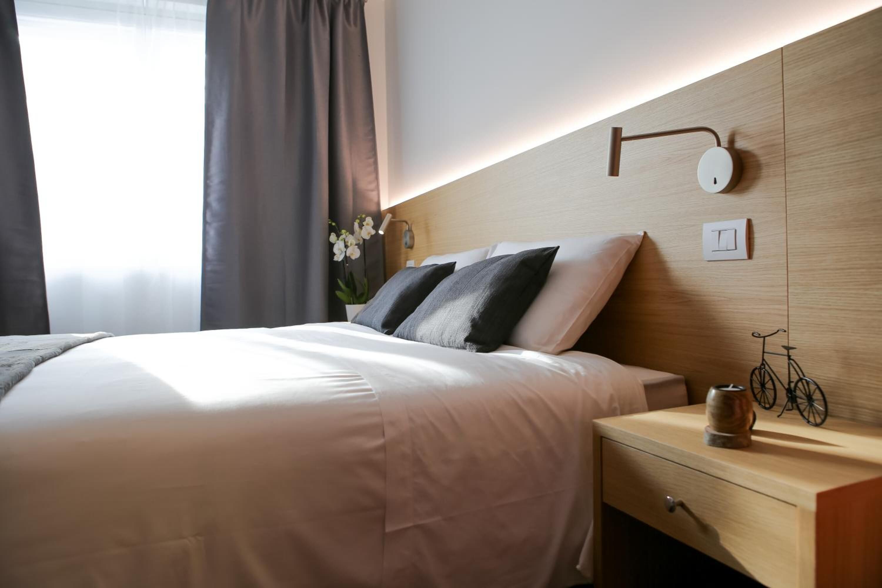 A modern hotel room with a neatly made bed, a bedside table with a lamp, and a wall-mounted light fixture.