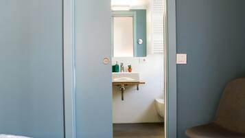 Un dormitorio con cama, una silla y una puerta que da a un baño con lavabo y espejo.