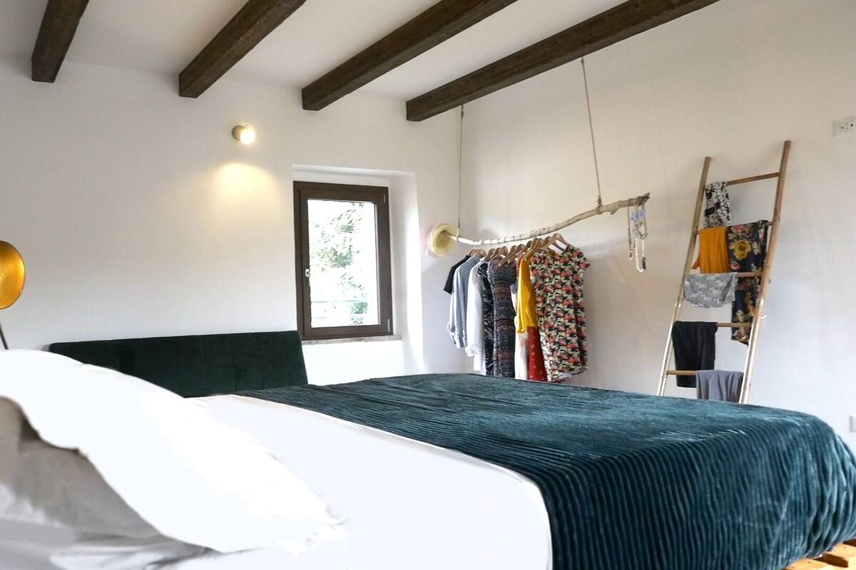 A bedroom with a bed, a hanging light, a window, and a clothes drying rack.